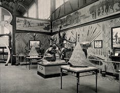 A sculpture of a man drumming while another one holds up a spear ready to attack another man who is lying on the ground. They are in the centre of a room that has knives and swords on display on the walls